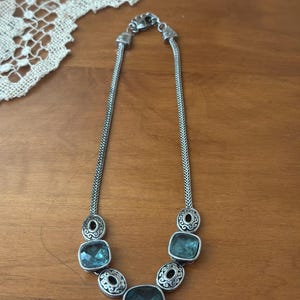 May include: Silver-toned necklace featuring a series of square, teal-colored gemstones. The necklace has a chain-like design with decorative circular accents between the gemstones. The necklace is displayed on a wooden surface.