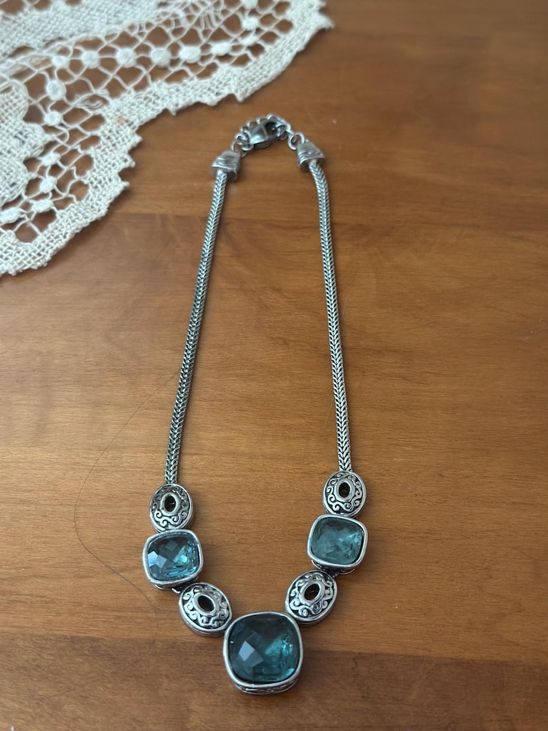 May include: Silver-toned necklace featuring a series of square, teal-colored gemstones. The necklace has a chain-like design with decorative circular accents between the gemstones. The necklace is displayed on a wooden surface.