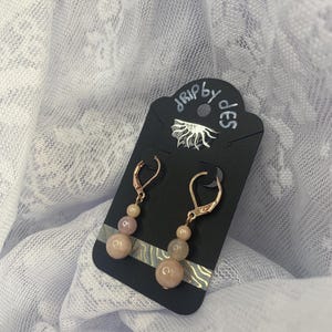 May include: A pair of dangle earrings with peach-colored beads and gold-colored ear wires. The earrings are displayed on a black card with the text "drip by des" in white script. The background is a white, lacy fabric.