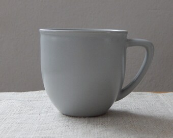 Puede incluir: Una taza de cerámica de color gris claro con forma redondeada y asa.