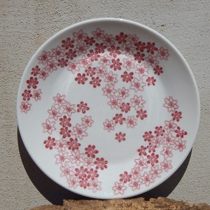 May include: A white ceramic plate with a pink floral pattern. The plate has a slightly raised rim and is decorated with a repeating pattern of small pink flowers.