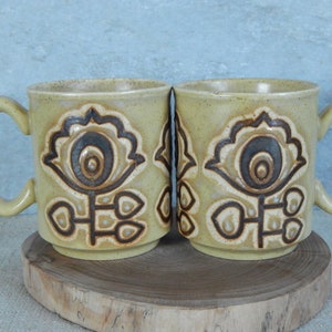 May include: Two yellow ceramic mugs with brown floral designs. The mugs have a rounded shape and a handle on the side.