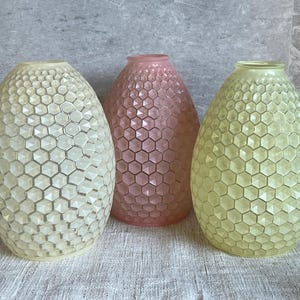 May include: Three decorative glass vases in a row. The vases are egg-shaped with a honeycomb pattern. One is off-white, one is pink, and one is light green. They sit on a white linen cloth.
