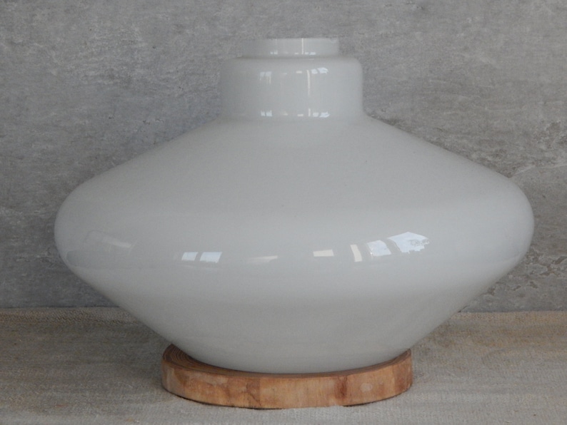 May include: White glass lampshade with a wide, rounded base and a narrow neck. The lampshade is sitting on a wooden base.