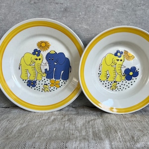以下が含まれることがあります： 黄色の縁取りと白い中心を持つセラミック製のプレートが2枚。プレートには、花と太陽が描かれた象の漫画のデザインが施されています。1枚のプレートには青い象と黄色い象が描かれており、もう1枚のプレートには2頭の黄色い象が描かれています。
