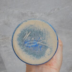 May include: The base of a blue ceramic vase with a white signature that reads "Nourie" in cursive.