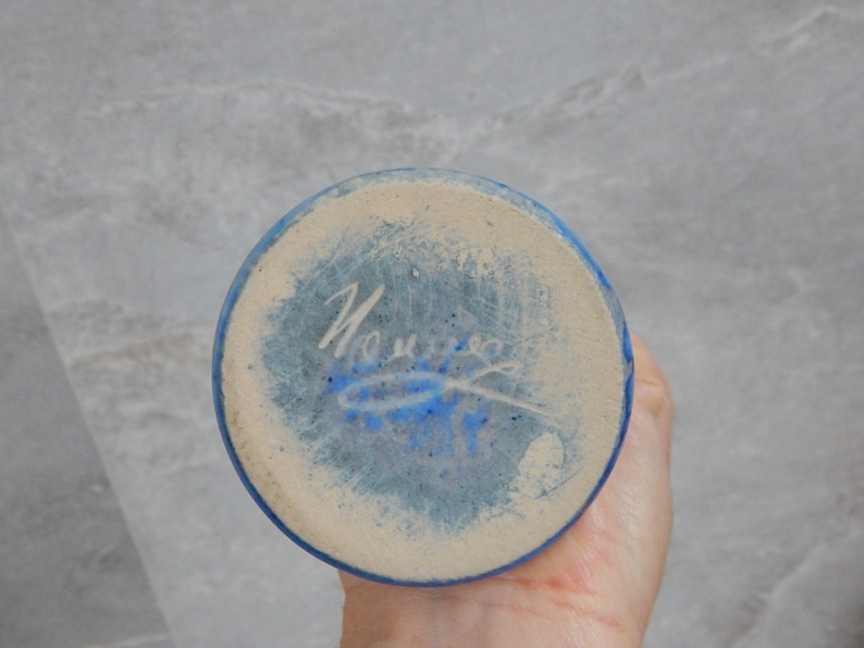 May include: The base of a blue ceramic vase with a white signature that reads "Nourie" in cursive.