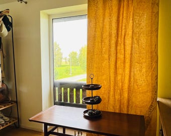 May include: A rectangular wooden table with tapered legs stands in front of a window covered by a long, mustard-yellow curtain featuring a subtle leaf pattern. A three-tiered dark metal serving stand sits on the center of the table surface.