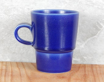 May include: A blue ceramic mug with a rounded handle. The mug has a slightly flared top and a smooth, glossy finish.