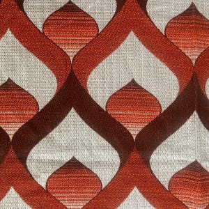 May include: Close-up of a fabric with a repeating pattern. The design features a series of interconnected, teardrop-shaped elements in shades of orange and brown against a neutral, textured background. The pattern creates a retro, geometric aesthetic.