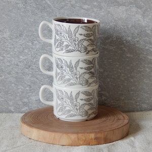 May include: A stack of four white ceramic mugs with a floral pattern in black and brown. The mugs are stacked on a wooden surface.