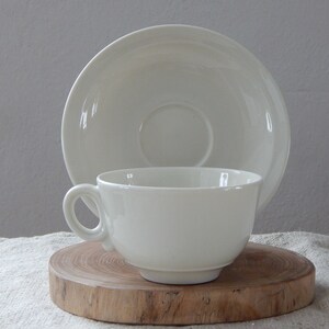 May include: A white porcelain teacup and saucer set. The cup has a rounded shape and a handle. The saucer is round and has a slightly raised edge.