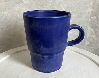May include: A cobalt blue ceramic mug with a textured surface and a rounded handle. The mug has a cylindrical shape with a stepped design near the base. The mug is sitting on a white surface.