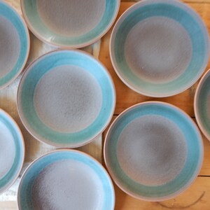 May include: Set of eight ceramic bowls with a light grey glaze and a teal blue rim. The bowls are arranged in a circle on a wooden surface.