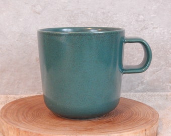 Peut inclure: Une tasse en céramique vert sarcelle avec une poignée arrondie. La tasse est posée sur un dessous de verre en bois.