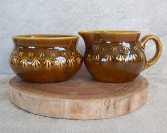 May include: A brown ceramic sugar bowl and creamer set with a decorative leaf pattern. The set is on a wooden base.