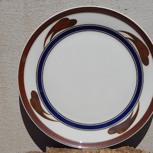 May include: A white ceramic dinner plate with a brown and blue design. The design features stylized brown leaves with a blue and white border.