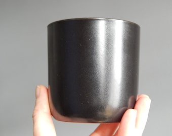 Puede incluir: Una taza de cerámica de color carbón oscuro con forma cilíndrica. La taza tiene una superficie lisa, ligeramente texturizada y un diseño simple y moderno. La taza está sujeta por una mano.