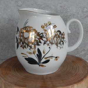 May include: White ceramic milk jug with a gold and black floral design. The jug has a rounded body and a handle.