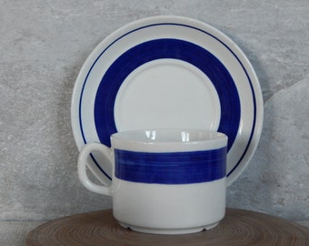 May include: White ceramic teacup and saucer set with a bold blue stripe design. The cup has a handle and sits on a matching saucer. The set is perfect for serving tea or coffee.