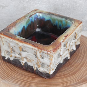May include: A small, square ceramic dish with a textured, white glaze and a dark brown rim. The inside of the dish is glazed in a blue, green, and brown colour.