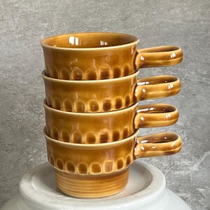 May include: A stack of four matching ceramic mugs in a glossy amber-brown glaze. Each mug features a rounded handle and a decorative band of vertical, arched indentations around the middle section, creating a textured, repeating pattern.