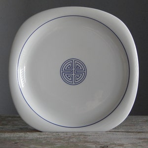 May include: White ceramic plate with a blue outline and a blue stylized symbol in the centre.