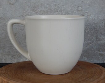 Peut inclure: Une tasse en céramique blanche unie de forme arrondie avec une anse. La tasse est posée sur une surface en bois.