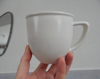 Puede incluir: Una taza de cerámica blanca con forma redondeada y asa. La taza se sostiene en una mano.