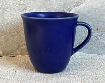 May include: A solid navy blue ceramic mug with a curved handle. The mug has a smooth, matte finish and a slightly flared rim. The mug is sitting on a textured, off-white surface.