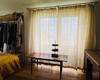 Puede incluir: Una mesa de centro rectangular de madera con patas cónicas se encuentra frente a cortinas de color claro, de largo hasta el suelo y con rayas verticales. Un soporte de metal de tres niveles descansa sobre la mesa.