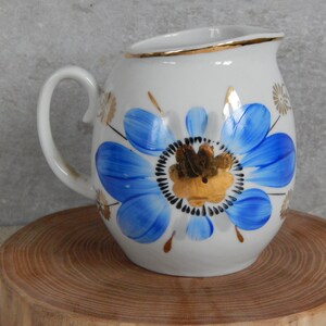 Può includere: Un creamer in ceramica bianca con un design floreale blu e oro. Il creamer ha un corpo arrotondato e un manico.