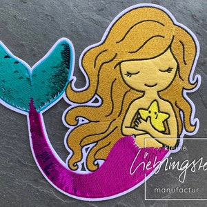 Può includere: Una sirena rosa e turchese con i capelli biondi che tiene una stella gialla. La sirena è ricamata con paillettes. Il testo "kleine liebingsteile manufactur" è in basso nell'immagine.