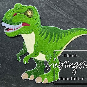 Könnte beinhalten: Grüne Filz-Dinosaurier-Applikation mit einem zahnigen Grinsen. Die Applikation ist perfekt, um Bastelarbeiten und Projekten eine verspielte Note zu verleihen. Der Text "kleine lieblingsteile manufactur" ist in der unteren rechten Ecke des Bildes sichtbar.