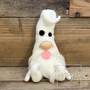 Könnte beinhalten: Eine weiße Zwergenpuppe mit langem, flauschigem Bart und spitzem Hut. Der Zwerg hat schwarze Augen, eine beige Nase und eine rosa Zunge, die herausragt. Die Puppe sitzt auf einer Holzoberfläche.