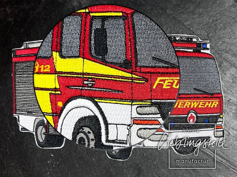 May include: Embroidered patch of a red and yellow fire truck with the word "FEUERWEHR" on the side. The patch is on a black background.