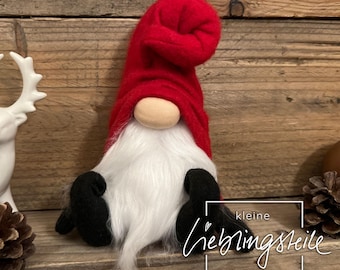 May include: A red and white gnome with a long white beard and black shoes. The gnome is sitting on a wooden surface. The text "kleine liebingsteile manufactur" is visible in the image.