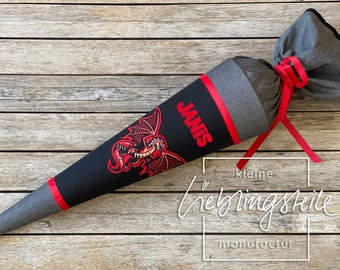 May include: A black and red school cone with a red dragon design and the name "JANIS" written in red. The cone is tied with a red ribbon and has a gray fabric base. The text "kleine lieblingsteile manufactur" is written in white on a black background.