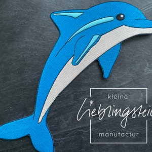 Może przedstawiać: Naszywka z haftowanego niebieskiego delfina z białym brzuchem. Naszywka pochodzi od firmy "kleine lieblingsteile manufactur".