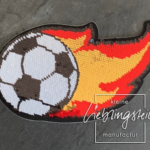 Pode incluir: Um remendo de lantejoulas preto, branco e vermelho de uma bola de futebol com chamas atrás. O remendo é feito por kleine lieblingsteile manufactur.