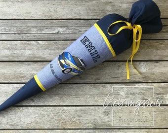 May include: A blue and gray fabric cone with a yellow ribbon and the name "EMIL" embroidered on it. A cartoon police car is printed on the cone. The text "13.08.2020" is printed on the cone. The cone is on a wooden surface.