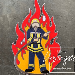 Può includere: Toppa ricamata da stirare con un vigile del fuoco che tiene un tubo davanti a una fiamma rossa e gialla. La toppa è etichettata "kleine lieblingsteile manufactur".