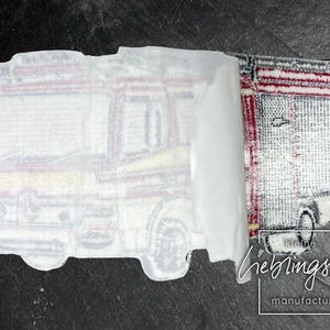 May include: A white iron-on patch with a detailed illustration of a fire truck. The patch is partially peeled back to reveal a red and white striped iron-on patch underneath. The text "kleine lieblingsteile manufactur" is visible in the bottom right corner.