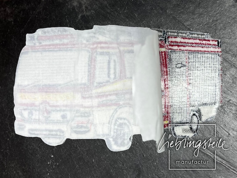 May include: A white iron-on patch with a detailed illustration of a fire truck. The patch is partially peeled back to reveal a red and white striped iron-on patch underneath. The text "kleine lieblingsteile manufactur" is visible in the bottom right corner.