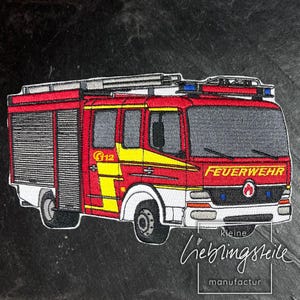 May include: Red and white embroidered patch of a fire truck with a ladder on top. The truck has the word "FEUERWEHR" on the front and the number "G12" on the side. The patch is from the company "kleine lieblingsteile manufactur".