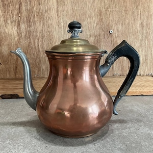 May include: A copper teapot with a brass lid and a black handle. The spout and the handle attachments are silver. The teapot has a rounded body and a decorative finial on the lid.