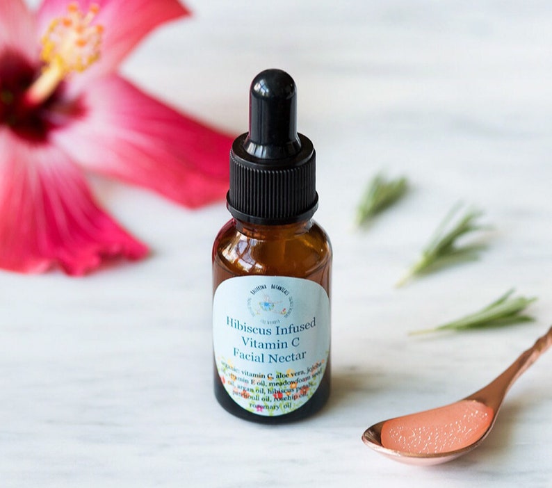May include: A small amber glass bottle with a black dropper top. The bottle is labeled "Hibiscus Infused Vitamin C Facial Nectar" and lists ingredients including vitamin C, aloe vera, jojoba oil, and hibiscus oil.