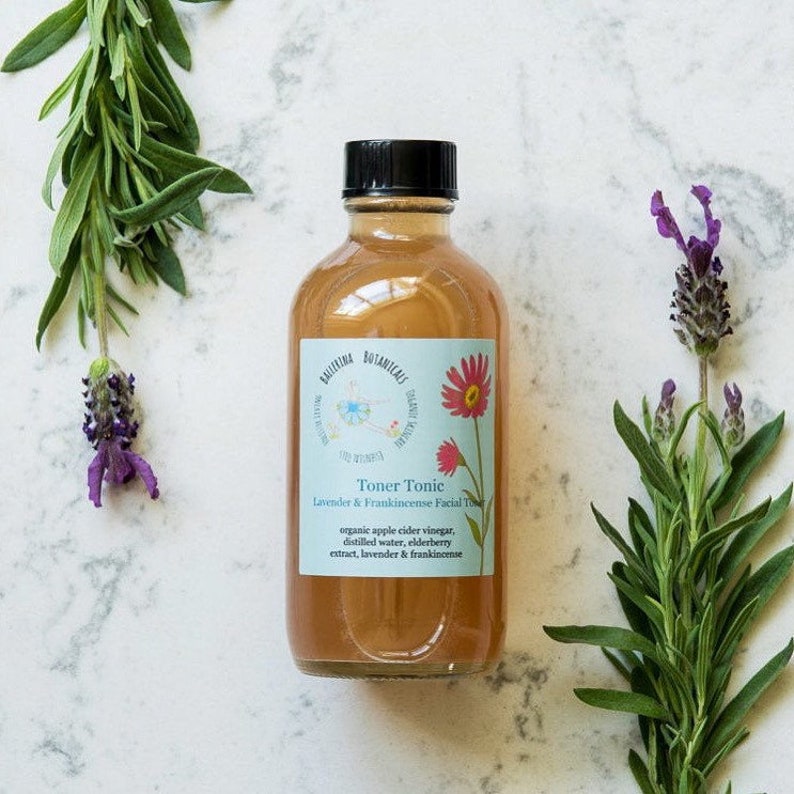 May include: A clear glass bottle with a brown liquid inside. The bottle has a black cap and a white label that says "Toner Tonic Lavender & Frankincense Facial Toner" and lists the ingredients: organic apple cider vinegar, distilled water, elderberry extract, lavender & frankincense.