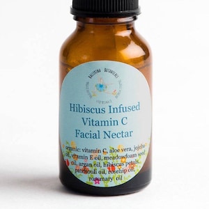 May include: A brown glass bottle with a blue and white label that reads "Hibiscus Infused Vitamin C Facial Nectar". The label lists the ingredients: organic vitamin C, aloe vera, jojoba oil, vitamin E oil, meadowfoam seed oil, argan oil, hibiscus petals, patchouli oil, rosehip oil, and rosemary oil.