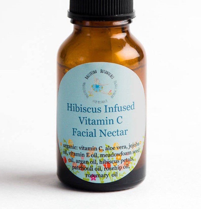 May include: A brown glass bottle with a blue and white label that reads "Hibiscus Infused Vitamin C Facial Nectar". The label lists the ingredients: organic vitamin C, aloe vera, jojoba oil, vitamin E oil, meadowfoam seed oil, argan oil, hibiscus petals, patchouli oil, rosehip oil, and rosemary oil.